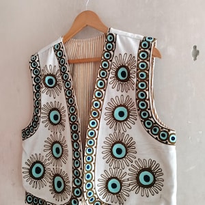 Op de afbeelding: Witte vest met een bloemmotief met turquoise en bruine accenten. Het mouwloze vest heeft een gestreepte voering en hangt aan een houten hanger tegen een witte muur.