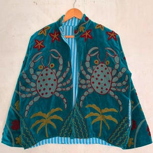Può includere: Una giacca in velluto color verde acqua con motivi di granchi ricamati, stelle marine rosse e palme dorate. La giacca presenta un colletto alla coreana, maniche lunghe e una chiusura frontale con zip, che rivela una fodera interna a righe verticali azzurre e bianche.