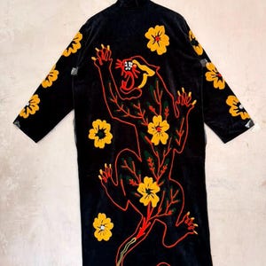 May include: A long black robe featuring a large embroidered panther outlined in red thread. The panther is surrounded by yellow flowers with red centres. The sleeves and body of the robe are decorated with additional yellow floral embroidery.