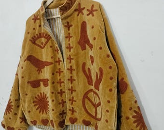 May include: Mustard-colored jacket with embroidered brown designs, including stars, hearts, and a peace symbol. The jacket has a round collar and a striped lining. The jacket is hanging on a wooden hanger.