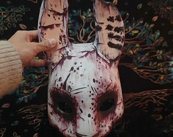 May include: A white rabbit mask with blood-like splatters and dark eye holes. The mask has long ears with brown and black markings. The mask is held by a person.