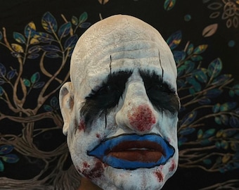 May include: A white clown mask with black eye makeup, red accents, and a blue mouth. The mask has a distressed appearance, with visible cracks and blood-like splatters. The mask is designed to be worn.