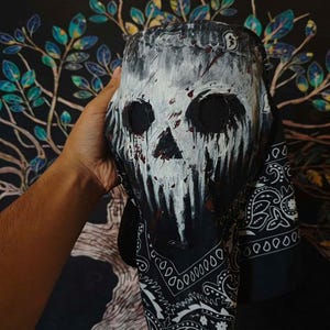Puede incluir: Una máscara de calavera en blanco y negro con pintura blanca goteando y detalles en rojo. La máscara está parcialmente cubierta por un pañuelo negro con un estampado de cachemira blanco. La máscara tiene cuencas de ojos grandes y un diseño de calavera detallado.