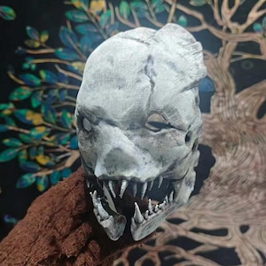 May include: A gray, monster-like mask with sharp teeth and empty eye sockets. The mask has a textured surface and is held against a brown, textured background. The mask's design suggests a horror or fantasy theme, suitable for cosplay or Halloween.