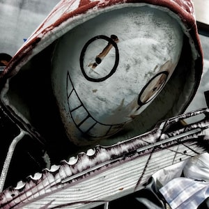 May include: A white mask with black eye and mouth details, and a red-trimmed hood. The mask has a circular eye design and a wide, toothy grin. The mask is holding a decorative object with a wing-like design.