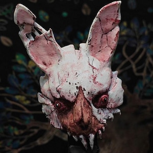 Op de afbeelding: Een wit en rood konijnenmasker met een gebarsten, verweerde uitstraling. Het masker heeft scherpe tanden, rode ogen en een dreigende uitdrukking. De oren zijn gebroken en het ontwerp suggereert een horrorthema.