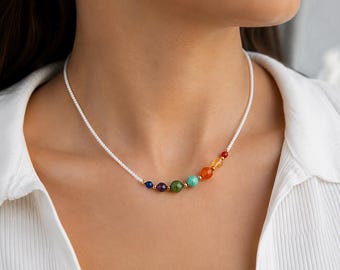 May include: A necklace featuring a white cord with a series of colorful gemstone beads. The beads are arranged in a gradient of colors, including blue, purple, green, orange, and red. The necklace is worn against a white shirt.