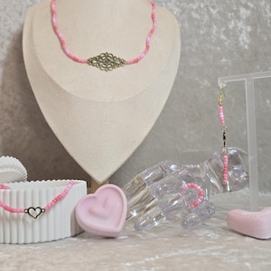 Könnte beinhalten: Ein Schmuckset bestehend aus einer rosa Perlenkette mit einem mittigen, antikgoldenen Filigrananhänger, einem passenden rosa Perlenarmband mit kleinem Herzanhänger und einem Paar langer, rosa Perlenohrringe mit herzförmigen Metallelementen.