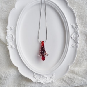 May include: A red glass pendant necklace with a silver chain. The pendant is shaped like a mushroom with white accents and a teardrop-shaped bottom. The necklace is displayed on a white decorative tray.