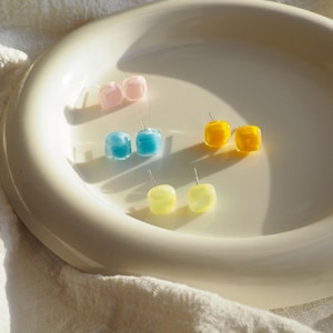 May include: A collection of colorful cube-shaped stud earrings in pink, blue, yellow, and light yellow. The earrings are displayed on a cream-colored plate, with a soft, textured background.