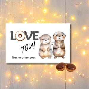 Pode incluir: Um cartão branco com as palavras "LOVE you!" e a frase "like no other one". Duas lontras em aquarela são apresentadas no cartão. A letra "O" em "LOVE" é um donut. Dois doces de chocolate na parte inferior.