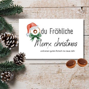 Może przedstawiać: Biała kartka świąteczna z napisem "du Fröhliche Merry christmas" i ilustracją czapki Świętego Mikołaja. Kartka leży na drewnianej powierzchni z igłami sosnowymi, szyszkami i dwoma cukierkami.