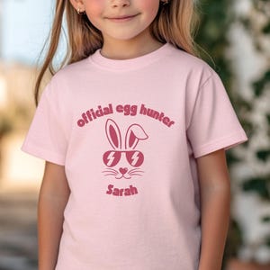 May include: A light pink t-shirt featuring the text "official egg hunter" in a dark pink font. Below the text is a graphic of a bunny wearing sunglasses, and the name "Sarah" is printed below the bunny.