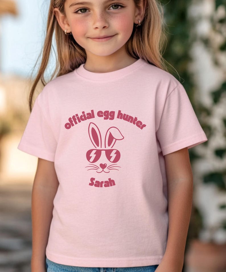 May include: A light pink t-shirt featuring the text "official egg hunter" in a dark pink font. Below the text is a graphic of a bunny wearing sunglasses, and the name "Sarah" is printed below the bunny.