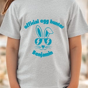 May include: A light gray t-shirt featuring the text "official egg hunter" above a cartoon bunny wearing sunglasses. The name "Benjamin" is printed below the bunny graphic in a matching blue color.