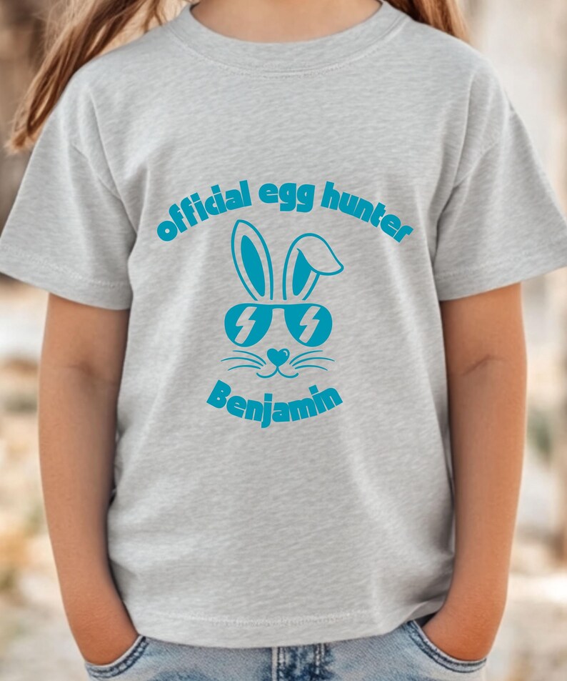 May include: A light gray t-shirt featuring the text "official egg hunter" above a cartoon bunny wearing sunglasses. The name "Benjamin" is printed below the bunny graphic in a matching blue color.