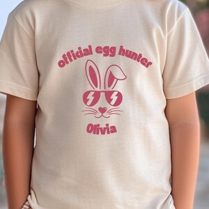 May include: A beige t-shirt featuring a pink graphic of a bunny wearing sunglasses with lightning bolt lenses. The text "official egg hunter" arches above the bunny, and the name "Olivia" is below.