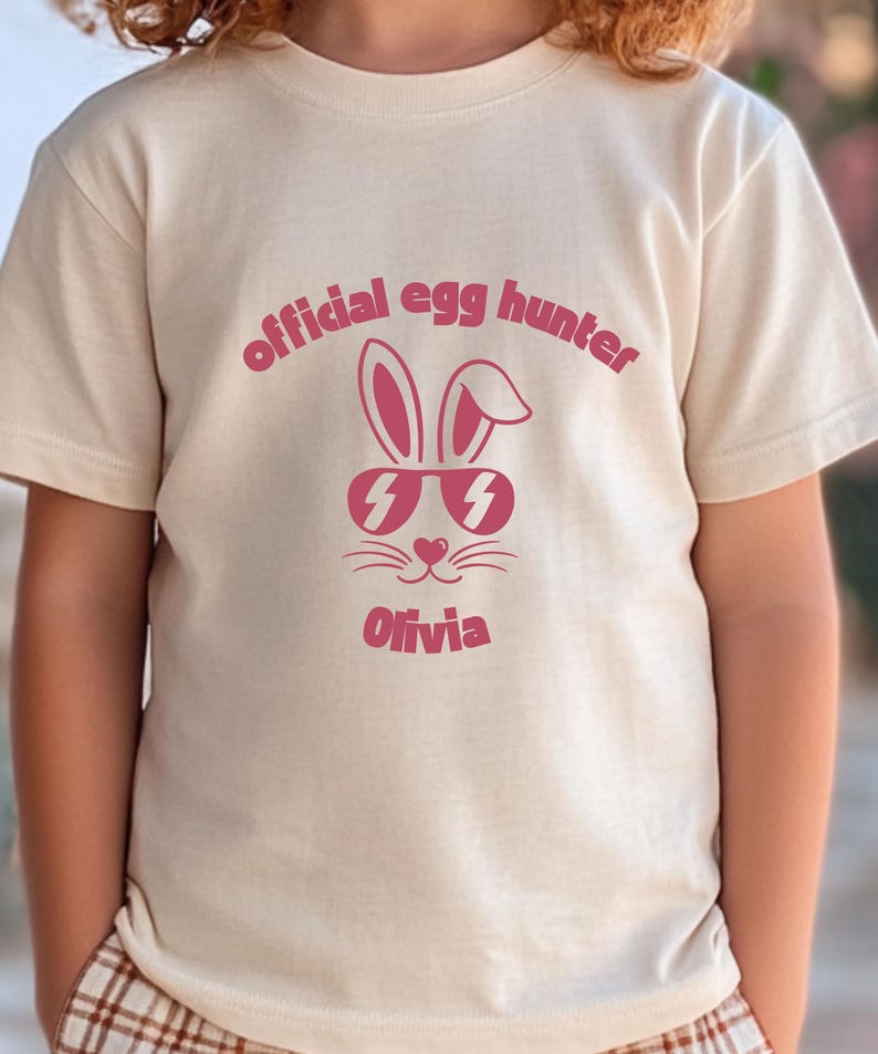 May include: A beige t-shirt featuring a pink graphic of a bunny wearing sunglasses with lightning bolt lenses. The text "official egg hunter" arches above the bunny, and the name "Olivia" is below.