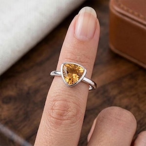 Könnte beinhalten: Ein silberner Ring mit einem dreieckigen, goldgelben Edelstein. Der Ring wird an einem Finger getragen, wobei das polierte Metallband und die leuchtende Farbe des Steins hervorgehoben werden. Der Ring ist ein Schmuckstück.