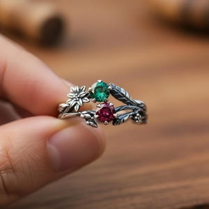 May include: A silver ring with a floral and leaf design, featuring two gemstones. One gemstone is a vibrant green, and the other is a deep red. The ring is held in a hand, showcasing the intricate details of the nature-inspired jewelry.