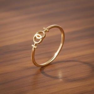 Può includere: Un anello dorato con due simboli femminili intrecciati. L'anello è posto su una superficie di legno calda, con un'ombra morbida. Il design dell'anello è semplice ed elegante.