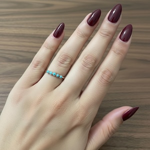 Op de afbeelding: Een delicate gouden ring met vijf kleine, ronde turkooizen stenen. De ring zit aan een hand met lange, diep bordeauxrode nagels. De achtergrond is een houten oppervlak.