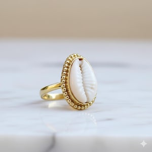Op de afbeelding: Een goudkleurige ring met een wit schelpmotief. De schelp is ovaal van vorm en gezet in een gouden frame met kralen. De band van de ring is van eenvoudig, gepolijst goud. De achtergrond is een zachte, neutrale tint.