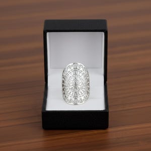 Pode incluir: Um anel prateado com um rosto oval e um design detalhado em filigrana. O anel é exibido dentro de uma caixa de joias preta com interior branco, sobre uma superfície de madeira castanha.