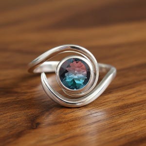 Puede incluir: Un anillo de plata con un diseño en espiral, con una piedra preciosa redonda con tonos turquesa y rosa. La banda del anillo se curva alrededor de la piedra, creando un aspecto único y elegante. El anillo está sobre una superficie de madera.