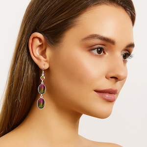 May include: Silver drop earrings with three colourful oval-shaped stones. The earrings feature a small, round, orange-toned stone at the top, followed by two larger, iridescent, oval stones with green, purple, and yellow hues. The earrings are a stylish accessory.