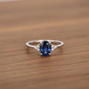 May include: A silver ring featuring a large, oval-cut, deep blue gemstone. Small, clear gemstones are set either side of the central stone. The ring is displayed on a warm, wooden surface, enhancing the ring's brilliance.