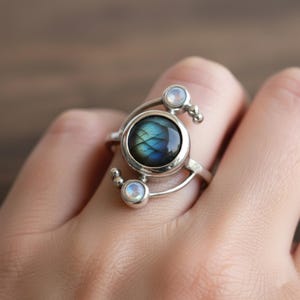 Può includere: Un anello d'argento con una pietra centrale rotonda blu-verde iridescente, circondata da pietre rotonde e chiare più piccole. L'anello ha un design artistico unico con dettagli argentati curvi.