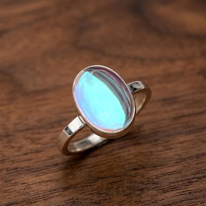 Puede incluir: Un anillo de plata con una piedra preciosa ovalada e iridiscente. La piedra preciosa tiene una mezcla de tonos azules, verdes y morados. El anillo está sobre una superficie de madera.