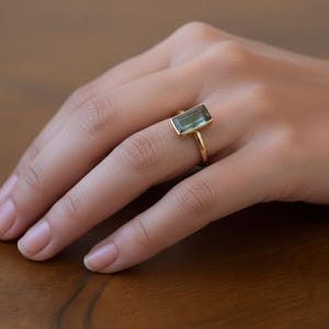 Może przedstawiać: Złoty pierścionek z prostokątnym, jasnozielonym kamieniem szlachetnym. Pierścionek ma prosty, elegancki design z cienką obrączką. Kamień szlachetny jest osadzony w złotej oprawie, tworząc minimalistyczną i nowoczesną estetykę. Pierścionek jest pokazany na dłoni.