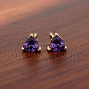 Könnte beinhalten: Ein Paar goldene Ohrstecker mit dreieckigen, tiefvioletten Edelsteinen. Jeder Ohrring hat eine goldene Fassung mit drei kleinen goldenen Akzenten um den Edelstein. Die Ohrringe liegen auf einer dunkelbraunen Holzoberfläche.