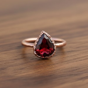 May include: A rose gold ring featuring a pear-shaped, deep red gemstone. The gemstone is framed by a textured, beaded border. The ring is set against a warm, wooden surface, highlighting the jewelry's design.
