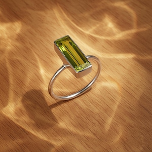 May include: A silver ring with a rectangular, light green gemstone. The gemstone is set in a rectangular bezel, and the band is a simple, thin silver. The ring is displayed on a wooden surface.