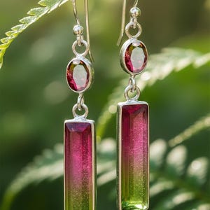 May include: Dangle earrings with a silver setting. Each earring has a small, oval, deep red gemstone at the top and a larger, rectangular gemstone below. The rectangular gemstones transition from red to green, creating an ombre effect.