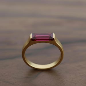 May include: A gold ring featuring a rectangular, deep red gemstone. The ring has a simple, modern design with a smooth, matte finish. The gemstone is set flush within the ring's setting, creating a sleek, elegant appearance.