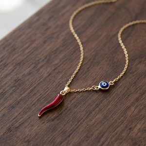 Op de afbeelding: Een goudkleurige ketting met twee hangers. Eén is een rode chilipeper, de andere een blauw en wit boze oog. De ketting wordt gepresenteerd op een houten oppervlak.