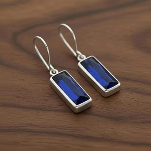 May include: A pair of silver-toned drop earrings with rectangular, deep blue gemstones. The earrings have a simple, elegant design, with a hook closure. The gemstones are set in a silver bezel, creating a polished look. The earrings are displayed on a wooden surface.
