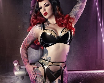 Puede incluir: Una mujer tatuada posa junto a una gran máquina de tatuar estilizada. Lleva un sujetador negro con lunares, liguero y medias. El fondo presenta luces de neón moradas y un entorno industrial futurista.