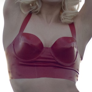Op de afbeelding: Een vrouw met blond haar en rode lippenstift draagt een rode latex bh met bandjes. Haar armen zijn boven haar hoofd geheven. De achtergrond is een heldere, bewolkte lucht.