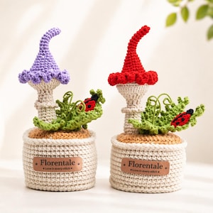 May include: Two crochet mushroom sculptures, one purple and one red, sit in beige crochet pots. Each has green leaves and a ladybug. The pots have a brown base and a leather tag that reads "Florentale. Love in every stitch."