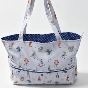 May include: A white tote bag featuring a repeating pattern of Peter Rabbit characters in various poses. The bag has two shoulder straps, a front pocket with a blue and white polka dot trim, and a matching blue and white polka dot interior lining.