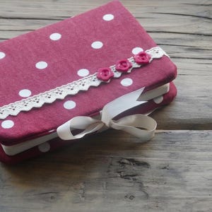 Può includere: Un diario rosso con pois bianchi, ornato da un bordo in pizzo bianco e tre bottoni rosa a forma di fiore. Un nastro color crema è legato attorno al diario, creando un fiocco decorativo.