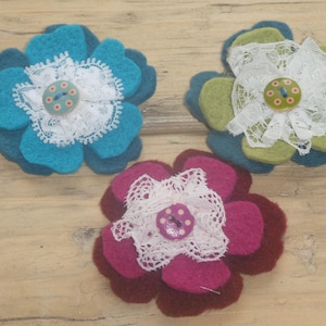 Può includere: Tre spille a forma di fiore in feltro fatte a mano, nei toni del blu, verde e bordeaux. Ogni fiore presenta un centro in pizzo bianco e un bottone. Le spille sono disposte su una superficie di legno marrone chiaro.