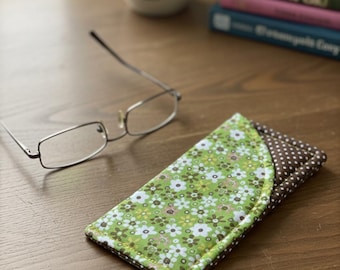 May include: A fabric glasses case with a green floral pattern on one side and brown polka dots on the other. The case is next to a pair of eyeglasses with a silver frame. A mug and books are in the background.