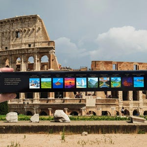 May include: A black box featuring landscape illustrations is held in front of the Colosseum in Rome. The box has Italian text. The illustrations show scenes such as mountains, sunsets, and water. The Colosseum is in the background.