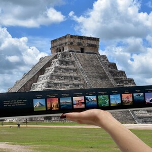 Puede incluir: Una mano sostiene un producto rectangular negro con temática de viajes frente a la pirámide de Chichén Itzá. El producto presenta imágenes de varios lugares emblemáticos. El cielo es azul con nubes blancas y esponjosas, y el suelo es hierba verde.